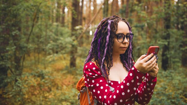 Donna in viaggio con i dreadlocks con un telefono nei boschi Viaggiatrice con zaino nei boschi che legge la mappa su uno smartphone e cerca una strada con tempo nuvoloso