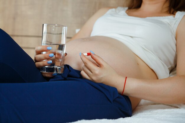 Donna incinta a letto con un bicchiere d'acqua e una capsula pillola il concetto di trattamento medico