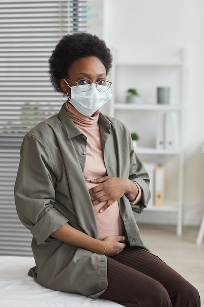 Donna incinta africana in maschera protettiva seduta sul divano in ospedale