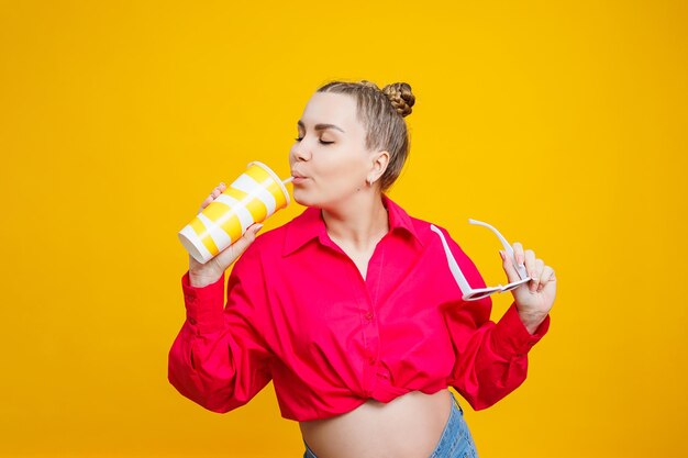 Donna incinta allegra in camicia rosa che tiene una bevanda gustosa su sfondo giallo isolato Una bevanda rinfrescante durante la gravidanza Una donna incinta beve acqua da un bicchiere usa e getta