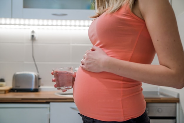Donna incinta bere acqua di acqua a casa in cucina. Felice donna incinta in possesso di un bicchiere di acqua, mentre in piedi in cucina