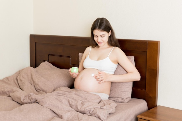 Donna incinta che applica una crema per smagliature sulla pancia, persone in gravidanza e concetto di maternità incinta, applica una crema antismagliature sulla pancia