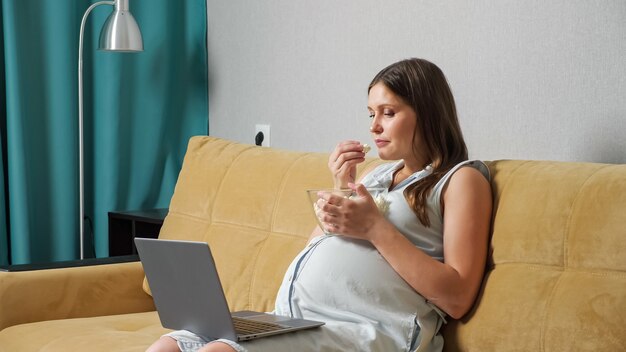 Donna incinta che guarda il laptop e mangia popcorn mentre è seduta sul divano