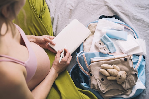 Donna incinta che impaccha la borsa dell'ospedale con la lista di controllo Madre incinta con una valigia di viaggio di vestiti per bambini che si prepara per la nascita del neonato che fa appunti nel diario