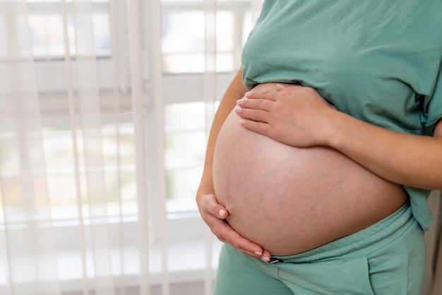 Donna incinta che indossa abiti comodi in piedi alla finestra Futura mamma che tiene il suo grande ventre con entrambe le mani Primo piano