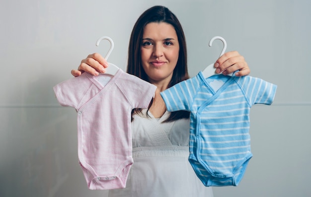 Donna incinta che mostra tute per bambini rosa e blu indoor. Messa a fuoco selettiva sui body in primo piano