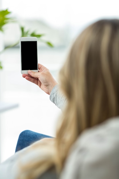 Donna incinta che si siede sul sofà e che per mezzo dello smartphone