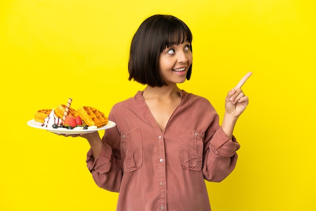 Donna incinta che tiene i waffle isolati su sfondo giallo che punta verso l'alto una grande idea