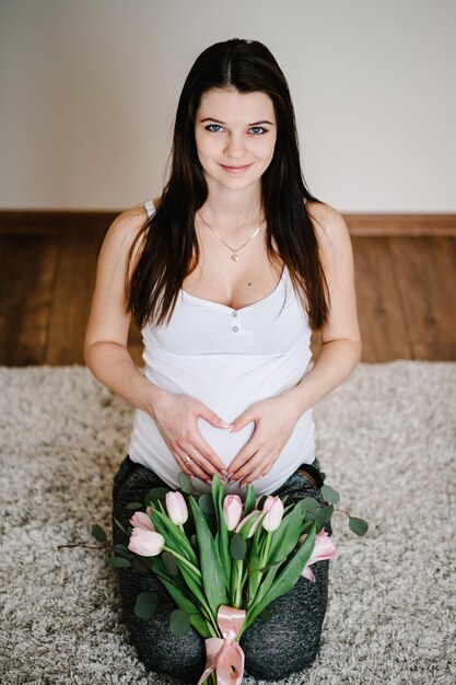 Donna incinta che tiene la pancia con un mazzo di fiori che abbraccia la pancia facendo il simbolo delle mani del cuore a casa Baby Shower Concetto di maternità Ragazza sulla foto