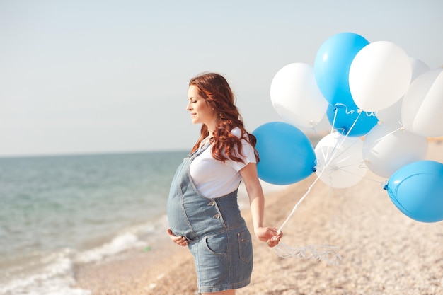 Donna incinta che tiene palloncini d'aria all'aperto. Camminando in riva al mare. Maternità.