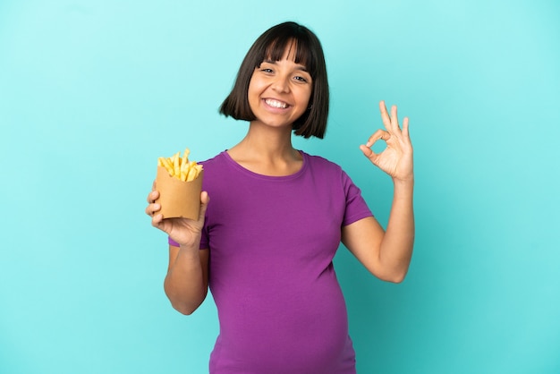 Donna incinta che tiene patatine fritte su sfondo isolato che mostra segno ok con le dita