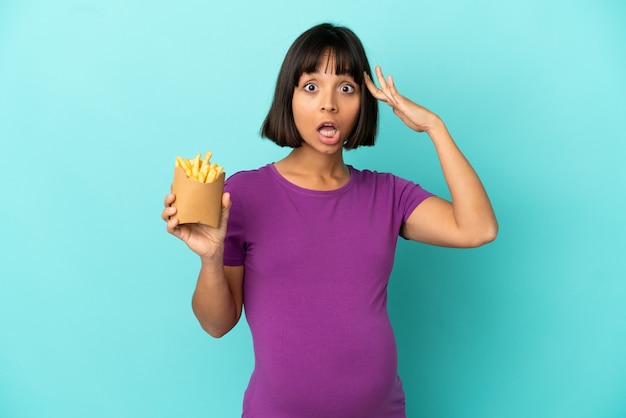 Donna incinta che tiene patatine fritte su sfondo isolato con espressione di sorpresa