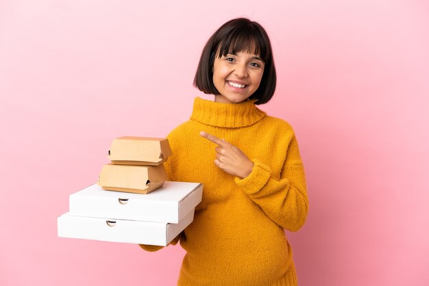 Donna incinta che tiene pizze e hamburger isolati su sfondo rosa che punta al lato per presentare un prodotto