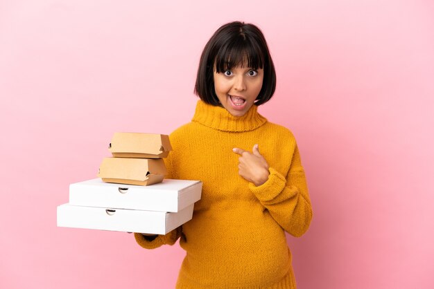 Donna incinta che tiene pizze e hamburger isolati su sfondo rosa con espressione facciale a sorpresa