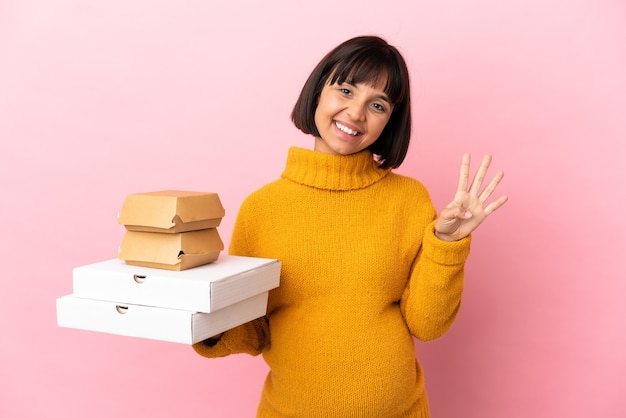 Donna incinta che tiene pizze e hamburger isolati su sfondo rosa felice e contando quattro con le dita