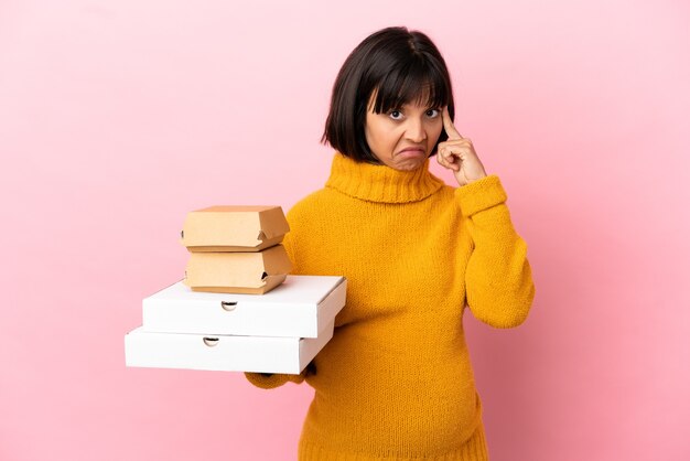 Donna incinta che tiene pizze e hamburger isolati su sfondo rosa pensando a un'idea