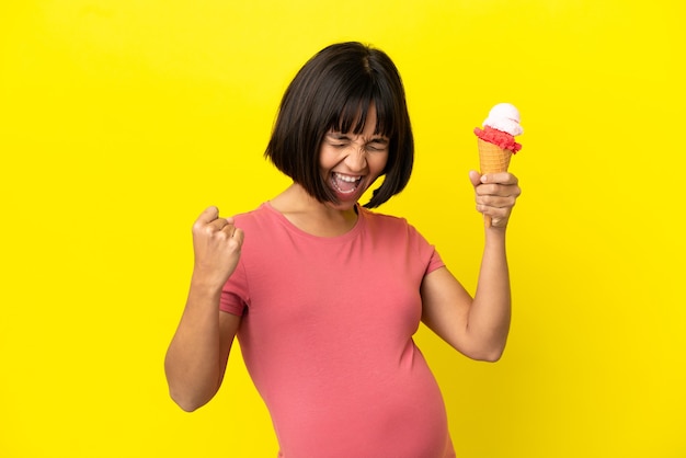 Donna incinta che tiene un gelato alla cornetta isolato su sfondo giallo che celebra una vittoria