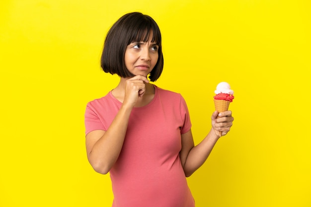 Donna incinta che tiene un gelato alla cornetta isolato su sfondo giallo che ha dubbi