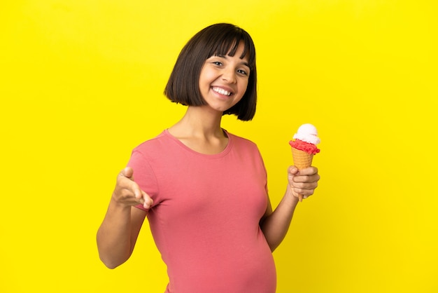 Donna incinta che tiene un gelato alla cornetta isolato su sfondo giallo che stringe la mano per chiudere un buon affare