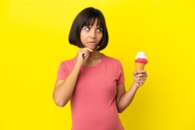 Donna incinta che tiene un gelato alla cornetta isolato su sfondo giallo con dubbi e pensieri