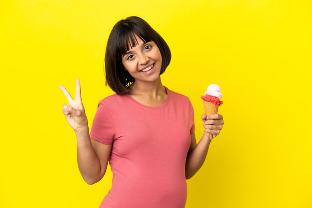 Donna incinta che tiene un gelato alla cornetta isolato su sfondo giallo sorridente e che mostra il segno della vittoria victory