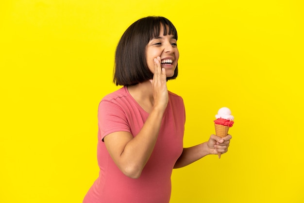 Donna incinta che tiene un gelato alla cornetta isolato sul muro giallo che grida con la bocca spalancata di lato