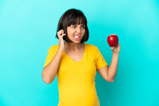 Donna incinta che tiene una mela isolata su sfondo blu frustrata e che copre le orecchie
