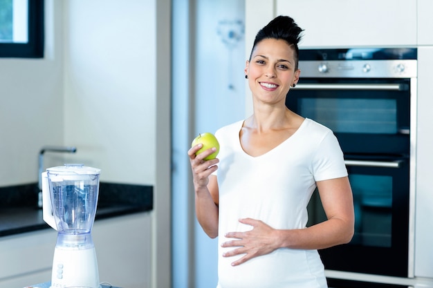 Donna incinta che tiene una mela verde e che sorride nella cucina