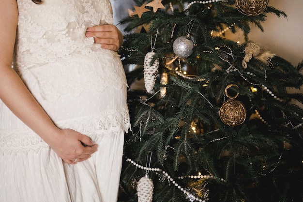 Donna incinta che tiene una pancia vicino a un albero di Natale. Abito bianco, primo piano della pancia.