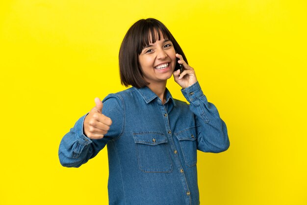 Donna incinta che utilizza il telefono cellulare isolato su sfondo giallo con il pollice in alto perché è successo qualcosa di buono