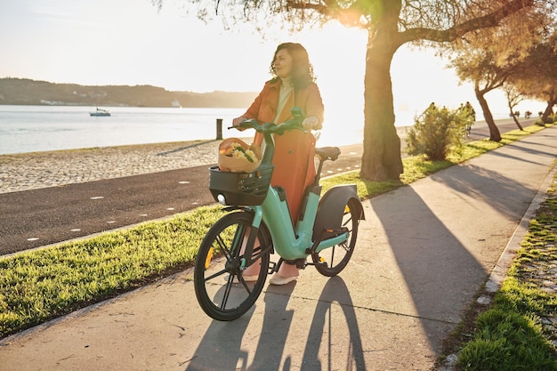 Donna incinta con cappotto arancione in bicicletta elettrica sul lungomare al tramonto