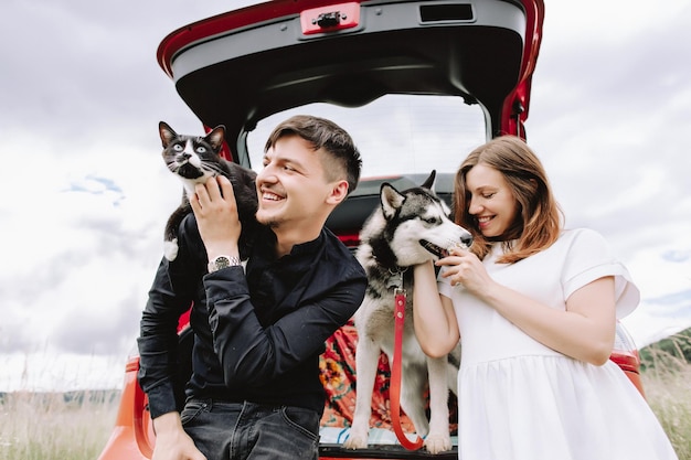 Donna incinta con husky e gatto sulle spalle dell'uomo vicino all'auto in auto sullo sfondo della natura