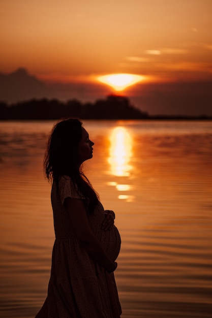 Donna incinta di profilo al tramonto vista laterale della silhouette nei raggi del sole al tramonto riflesso nell'acqua...