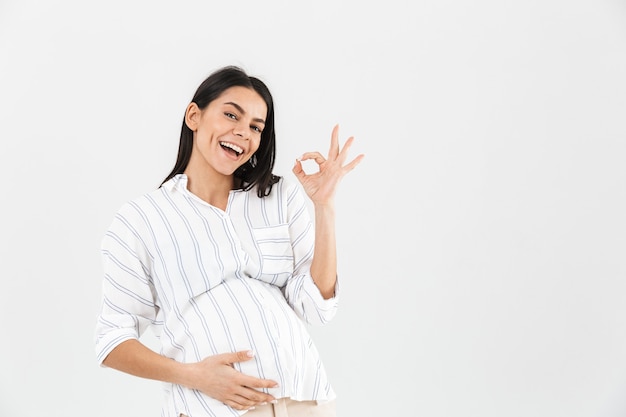 donna incinta felice con la grande pancia sorridente e mostrando segno giusto isolato sopra il muro bianco