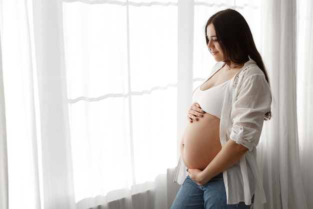Donna incinta in piedi vicino alla finestra in appartamento accarezza abbracciando la pancia pancia