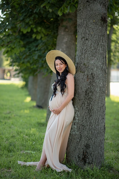 Donna incinta in posa in un abito beige con alberi verdi.