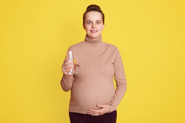 Donna incinta in posa isolata sopra il muro giallo, in piedi con un bicchiere d'acqua indossando abbigliamento casual, toccando la sua pancia con la mano.