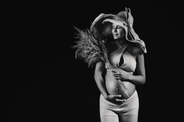Donna incinta in un cappello di paglia con abiti beige con un bouquet in mano in studio su sfondo nero