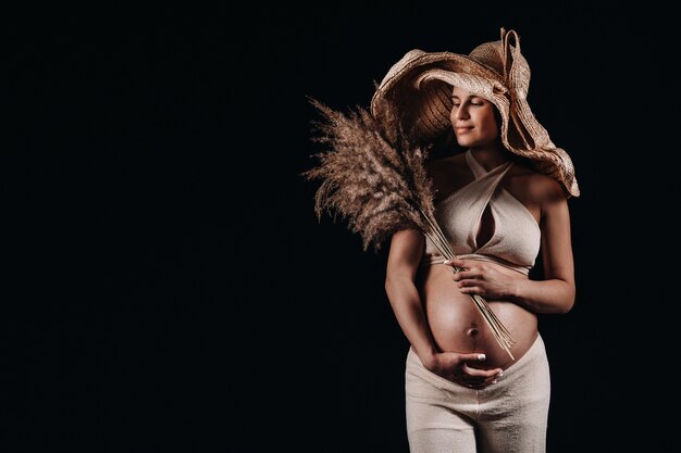 Donna incinta in un cappello di paglia con vestiti beige con un bouquet in mano in studio su sfondo nero.