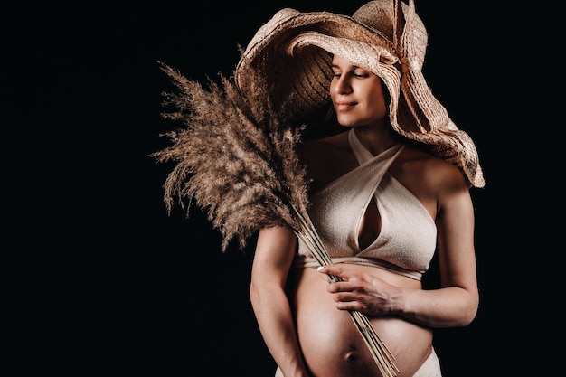 Donna incinta in un cappello di paglia con vestiti beige con un bouquet in mano in studio su sfondo nero.