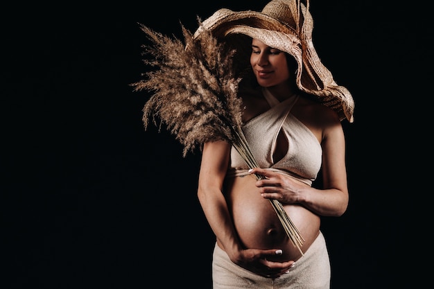 Donna incinta in un cappello di paglia con vestiti beige con un bouquet in mano in studio su sfondo nero.