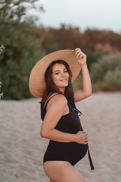 donna incinta in un costume da bagno nero e cappello su una spiaggia sabbiosa