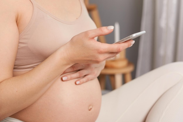 Donna incinta irriconoscibile seduta a letto con indosso un top beige utilizzando il telefono cellulare che digita messaggi navigando in internet tenendo lo smartphone