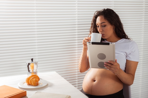 Donna incinta ispanica con tavoletta digitale durante la colazione. Tecnologia, gravidanza e maternità.