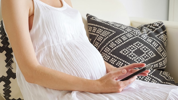 Donna incinta naviga in internet con lo smartphone seduto sul divano di casa primo piano