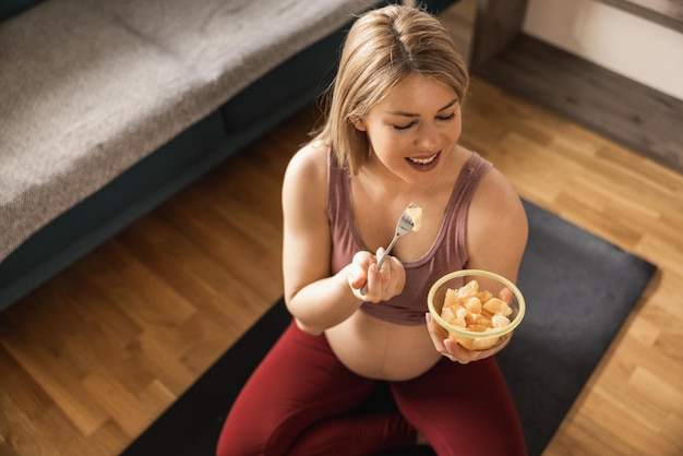 Donna incinta nel suo salotto con una sana colazione con frutta fresca dopo l'allenamento.