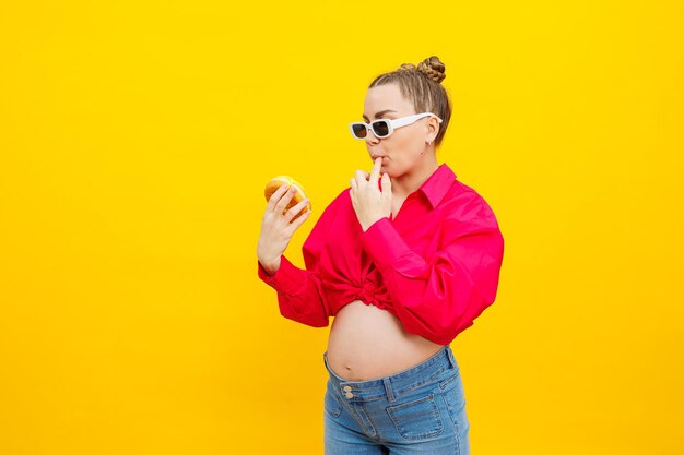 Donna incinta positiva che mangia ciambelle indossando camicia rosa isolata su sfondo giallo Felicità dalla gravidanza mentre si aspetta un bambino Foto di alta qualità Cibo nocivo durante la gravidanza