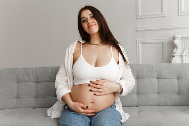 Donna incinta seduta nell'appartamento che abbraccia la pancia, l'addome e si diverte della gravidanza.