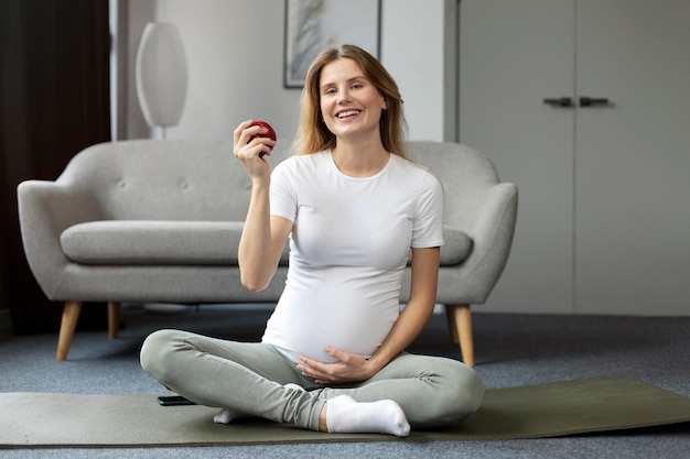 Donna incinta sorridente che pratica yoga nella posa del loto tenendo la mela rossa a casa Yoga in gravidanza