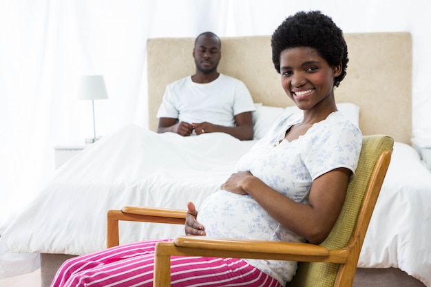 Donna incinta sorridente che si siede sulla sedia e sull'uomo che si siedono sul letto nel fondo